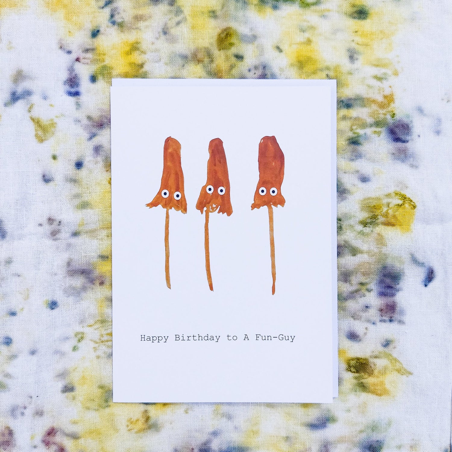 Birthday Cards. White card featuring three orange mushrooms with black and white eyes. Under the mushrooms it says ' Happy Birthday to A Fun-Guy'. Behind the card is a naturally dyed fabric background with yellow and warm blue tones. - Duck Egg Designs Co