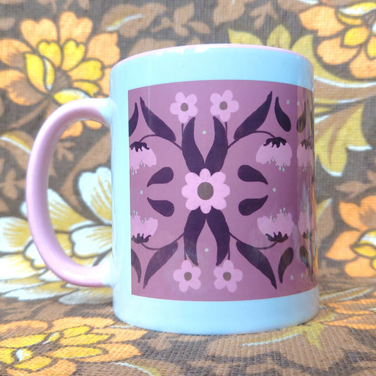 Pink and white mug with a symmetrical floral retro inspired pattern sits in front of a white and brown floral background. The mug features a pink handle and inside, with a border of white around the design.