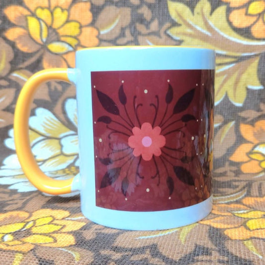 A white mug with a yellow handle and inside sits in front of a brown and yellow floral background. The mug features a brown rectangle across it with a dark orange and black floral pattern.