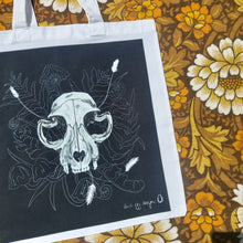 Load image into Gallery viewer, A close up of the white tote bag featuring a black square with a white cat skull featuring leafy fronds and fungi growing out from behind it. To the right of the bag you can see a floral patterned background in white, brown and yellow. 
