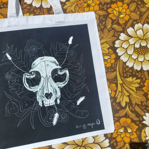 A close up of the white tote bag featuring a black square with a white cat skull featuring leafy fronds and fungi growing out from behind it. To the right of the bag you can see a floral patterned background in white, brown and yellow. 