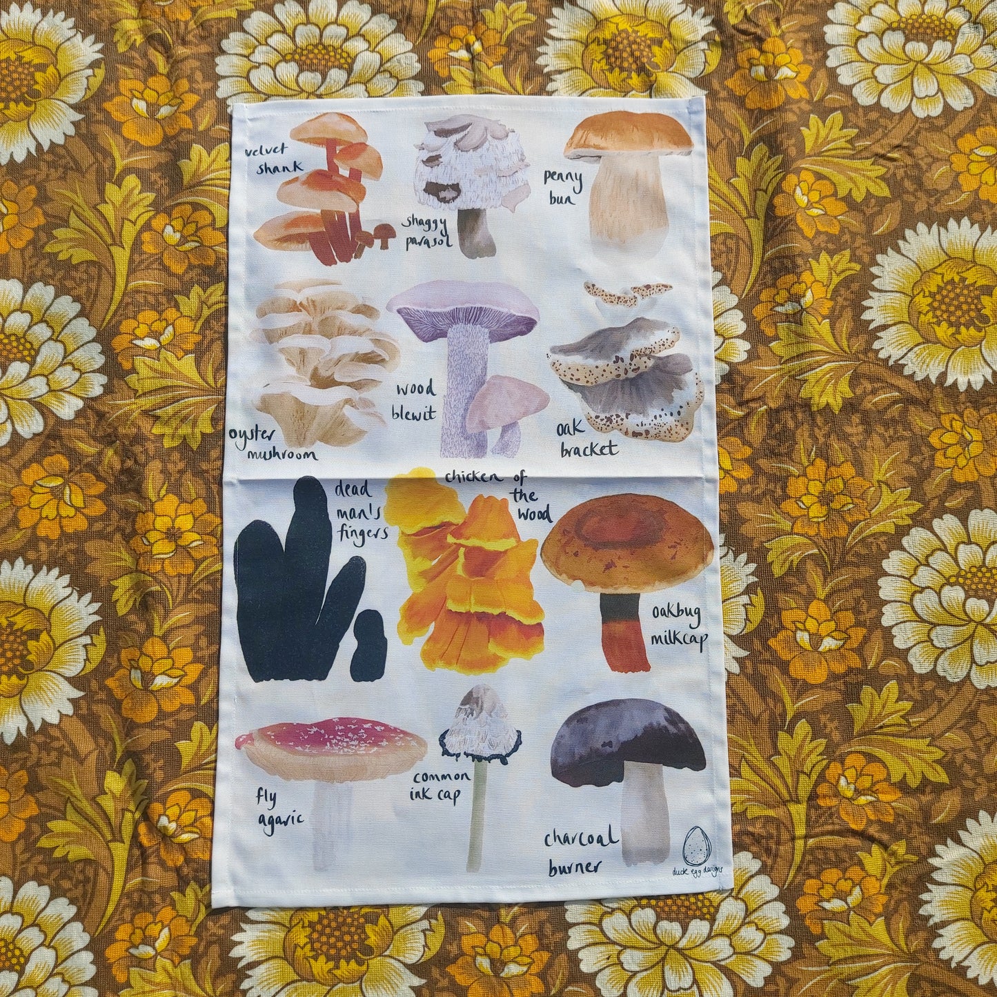 An off white tea towel featuring 12 different British fungi in rows of three with their names in black handwriting next to them. Behind the tea towel is a warm brown retro floral patterned fabric.