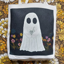 Load image into Gallery viewer, A close up of view of a white tote with a black square printed on it featuring a white ghost holding a grey kitten amid a row of pink, red and mustard yellow colours. Behind the tote you can see a warm brown floral retro patterned fabric.
