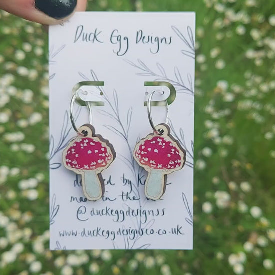 Silver hoops with red and white wooden toadstool charms on a white backing card in front of a sea of daisies.