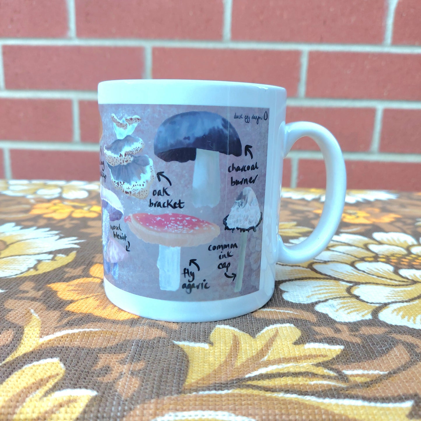 The right hand side of a white mug with a british fungi design sat on a warm brown retro floral patterned fabric in front of a red brick wall.