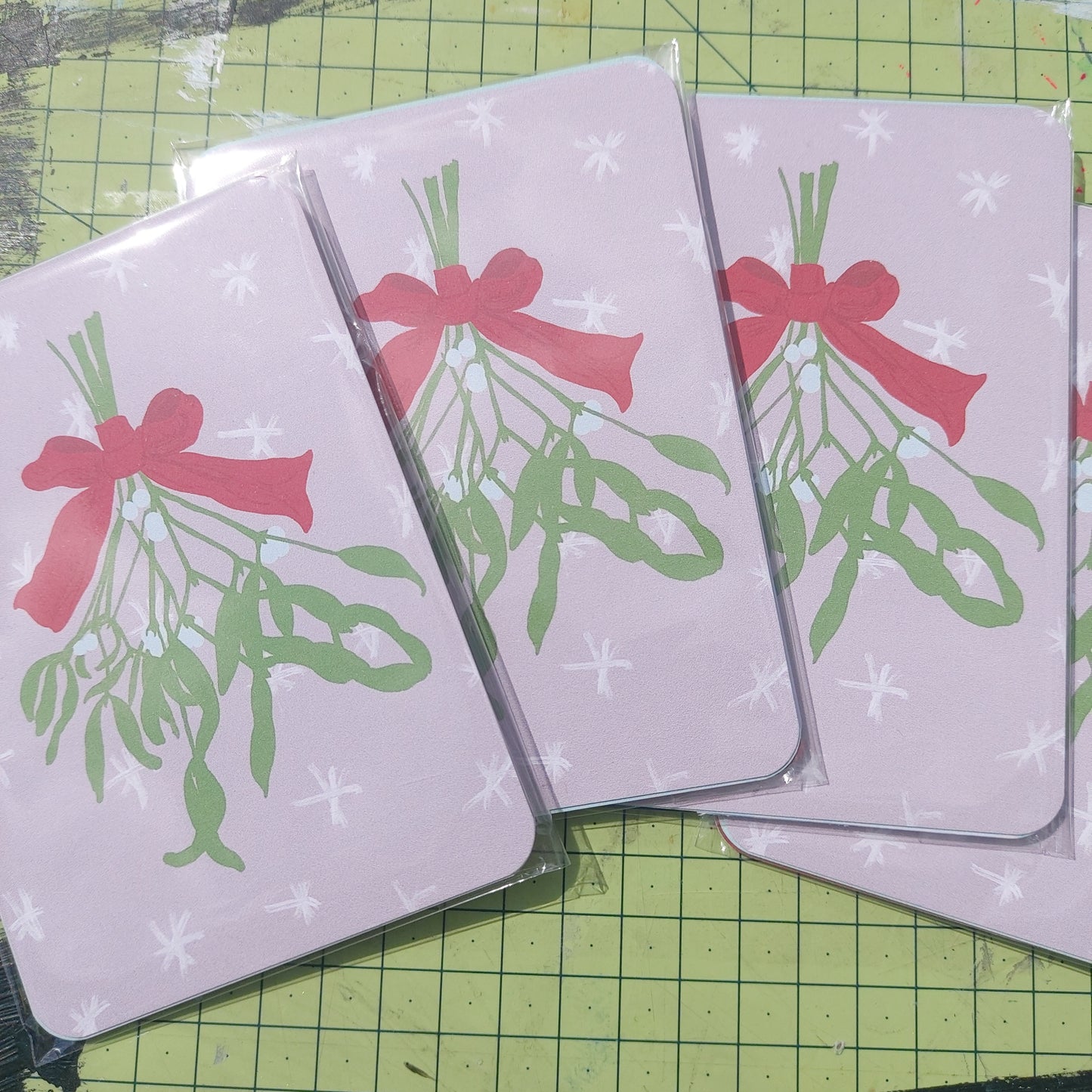 Four packs of Christmas postcards across a green cutting mat. You can see the top postcard in the pack which is a pale pink design featuring white snowflakes and a bunch of mistletoe tied by a red ribbon hanging down the middle.