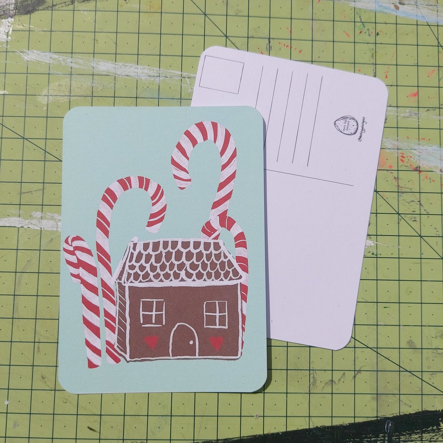 Front and back of a postcard are visible on a green cutting mat. Front of the postcard shows a gingerbread house with red and white icing under some tall red and white candy canes on a peppermint green background. The back of the postcard is white, featuring lines for the address and a square for the stamp.