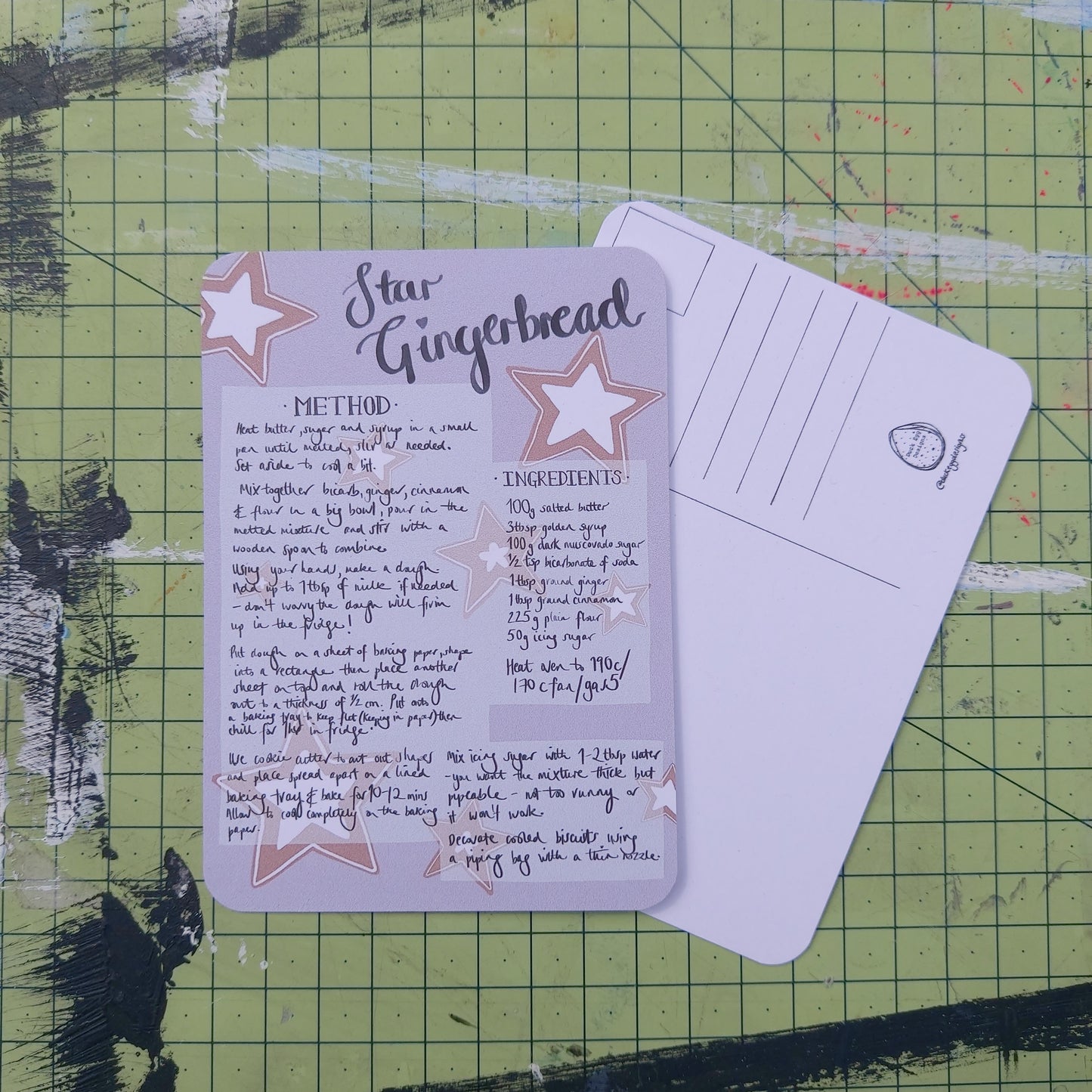 Front and back of a postcard are visible on a green cutting mat. The front is pale purple with a handwritten star gingerbread recipe. The back is white with lines for the address and a square for the stamp as well as the duck egg designs logo.