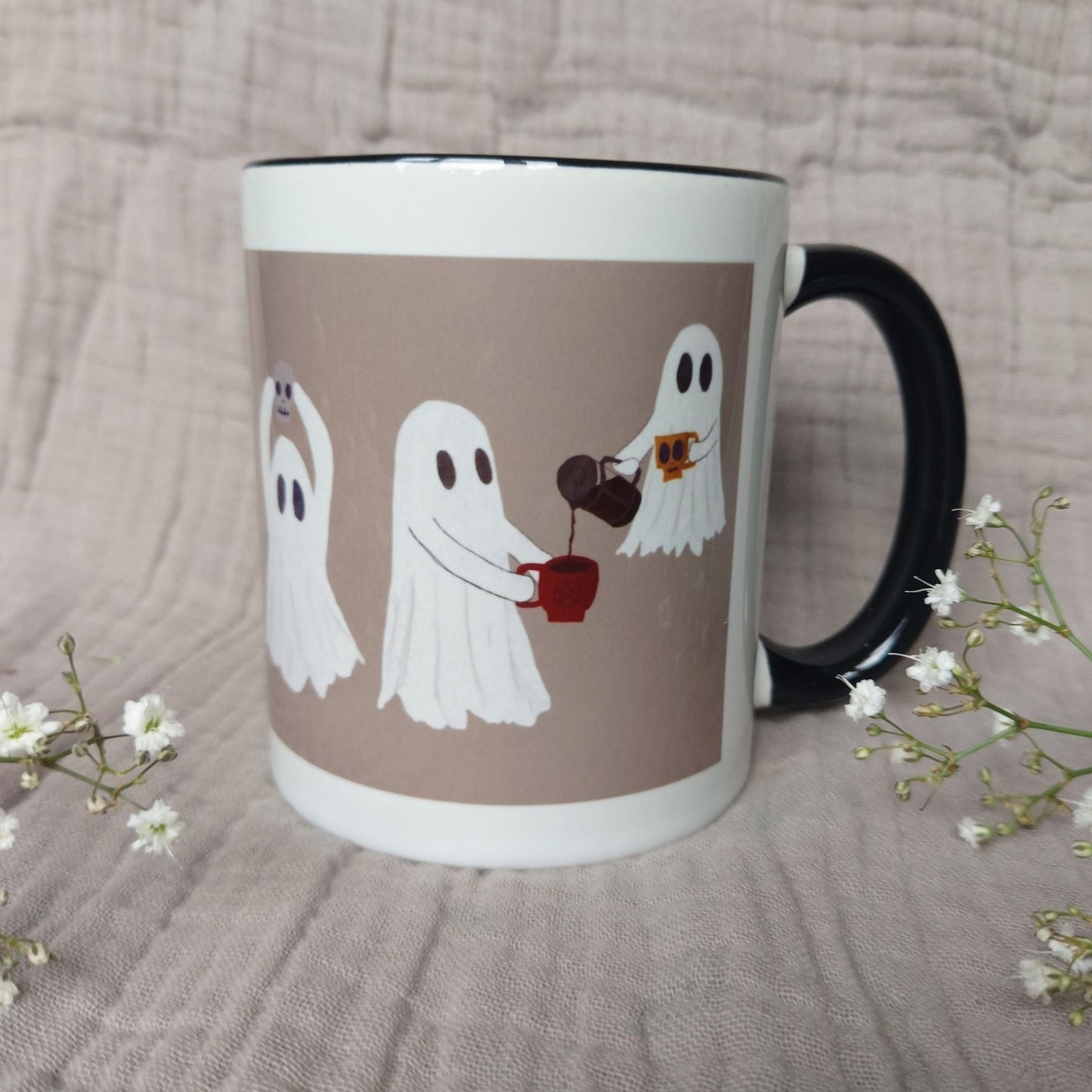 A white and warm grey mug featuring ghost with a black interior and handle sits in front of a greyish lilac material in between two sprigs of babys breath. The two ghosts you can see the most are sharing a pot of coffee - with one of the ghost floating above the other.