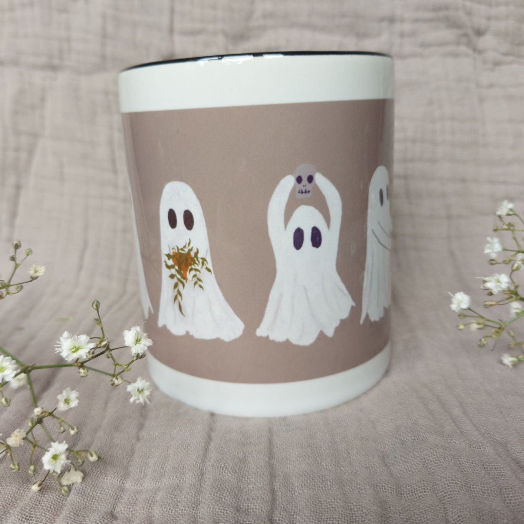 A white and warm grey mug featuring ghost with a black interior sits in front of a greyish lilac material in between two sprigs of babys breath. The two ghosts you can see clearly hold a plant pot with a green trailing plant, and a mini skull.