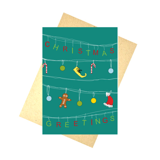 A blue card with white lines across it, from which the letters for the works 'Christmas Greetings' hand in alternating green and red letters. Hanging across the middle two rows are a mix of red and white candy canes, green, blue and yellow baubles, a yellow elf shoe and a santa hat. Behind the card is a brown envelope, behind which is a white background.